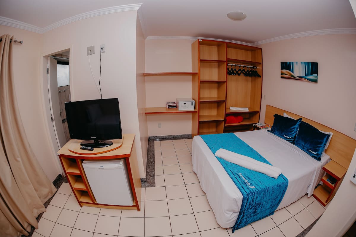A hotel room with a bed, a TV, a mini-fridge, a wardrobe, and a painting on the wall.