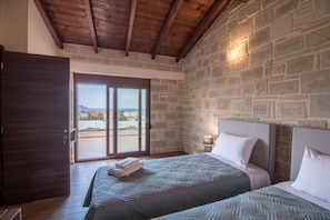 A bedroom with two beds, a stone wall, a wooden ceiling, and a view through a glass door.