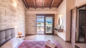 A bedroom with a stone wall, a wooden ceiling, a bed with a headboard, a nightstand, a lamp, a bathroom with a mirror, and a view of the sea through a glass door. - Villa Katerina (Chania)