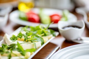 Une vue rapprochée d’un plat contenant du fromage et des herbes, un bol blanc rempli d’un liquide foncé, et une assiette avec des légumes coupés en tranches en arrière-plan.