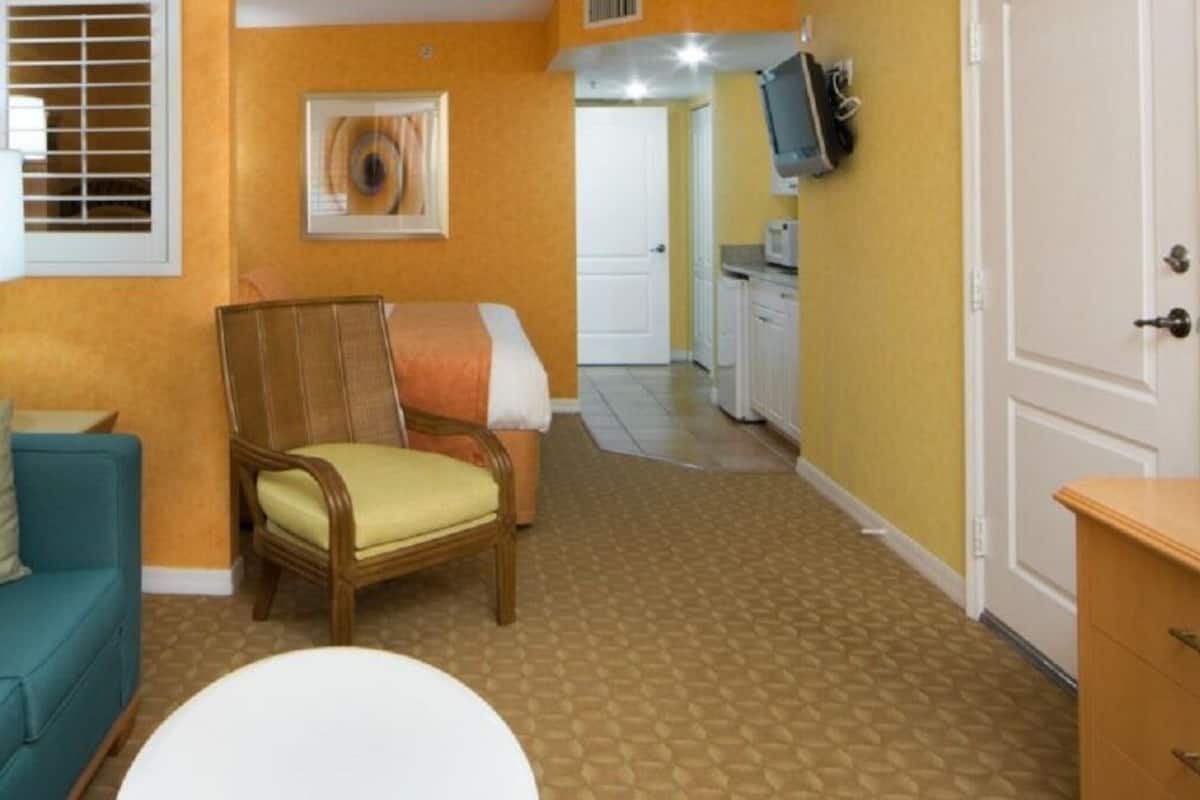 A compact living area with a teal sofa, a wooden chair, a white table, and a small white table. A painting is on the wall, and a television is mounted above a kitchen counter.