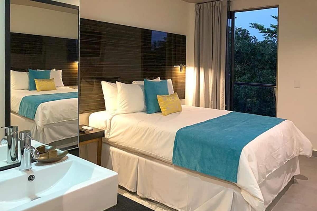Een moderne slaapkamer met een groot bed, een nachtkastje met spiegel, een witte wastafel en uitzicht op groen.