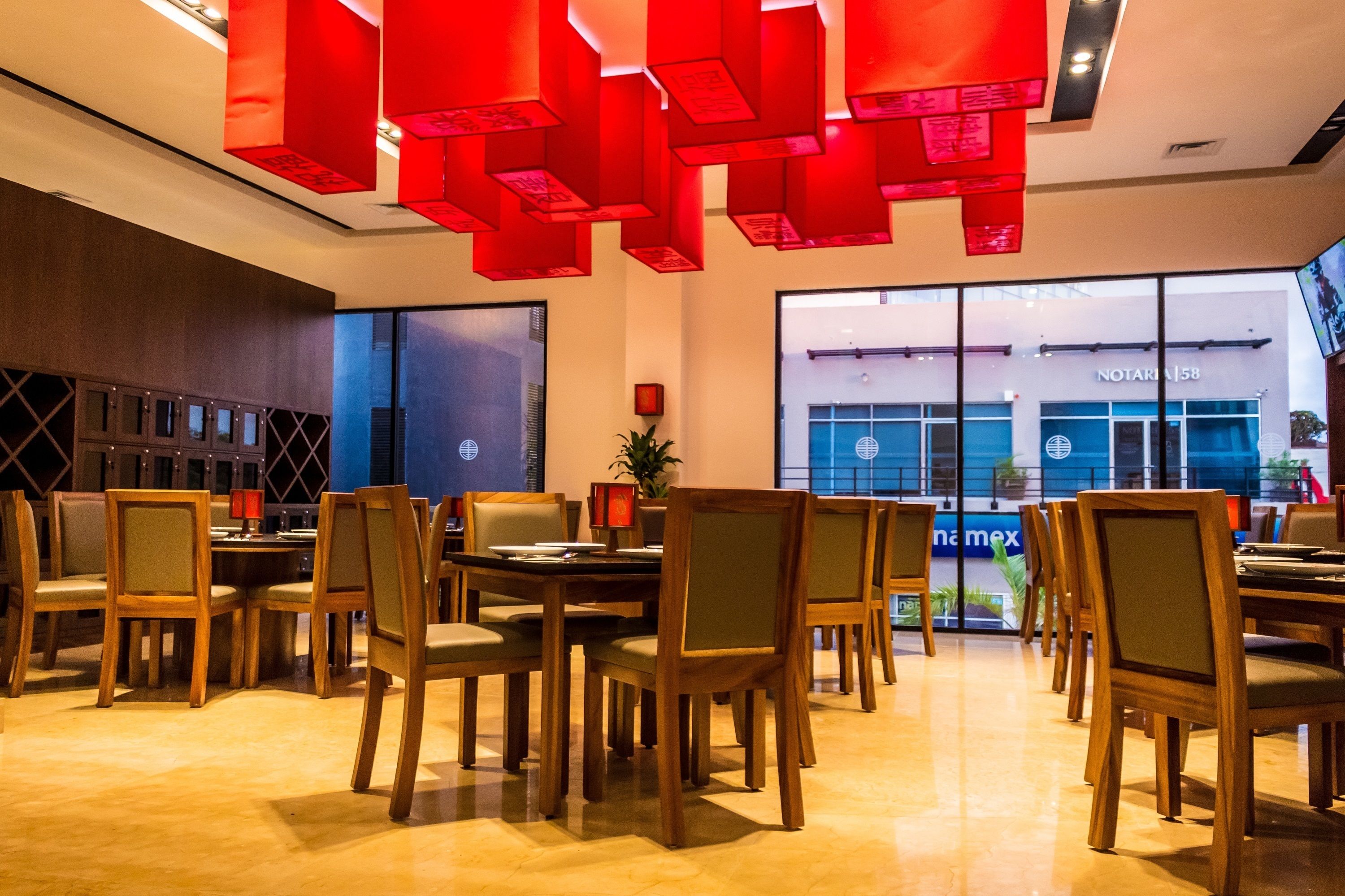 A modern dining area with wooden tables and chairs, red hanging lights, and large windows with views of the street.
