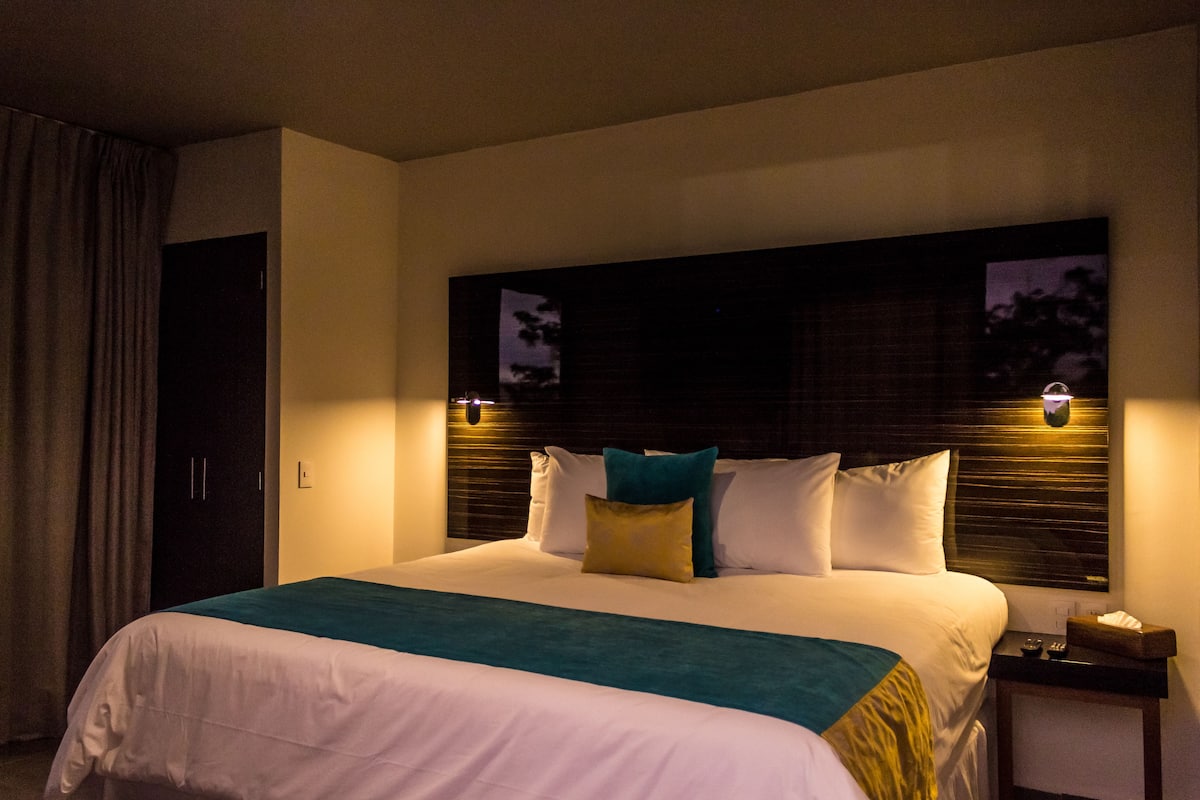 Hotelkamer met een groot bed, een hoofdbord met ingebouwde verlichting, een nachtkastje met lamp en een raam met uitzicht op bomen.