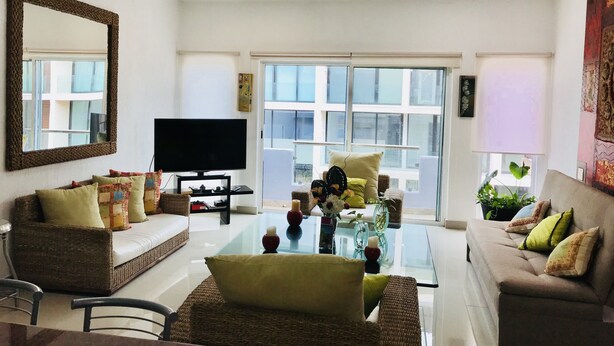 Un salon spacieux doté d’un grand téléviseur à écran plat, de meubles en osier, d’une table basse en verre et de grandes fenêtres offrant une vue sur la ville.