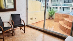 Une pièce avec une grande fenêtre, une chaise et une vue sur un bâtiment entouré d’une clôture blanche, avec une plante en pot.