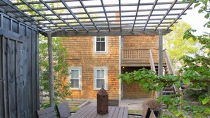 Une terrasse en bois avec une pergola, une table entourée de chaises et, au fond, un bâtiment doté de fenêtres.