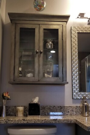 Une salle de bain avec un comptoir en marbre, un lavabo blanc, un meuble avec des portes en verre et un miroir doté d’un cadre texturé.