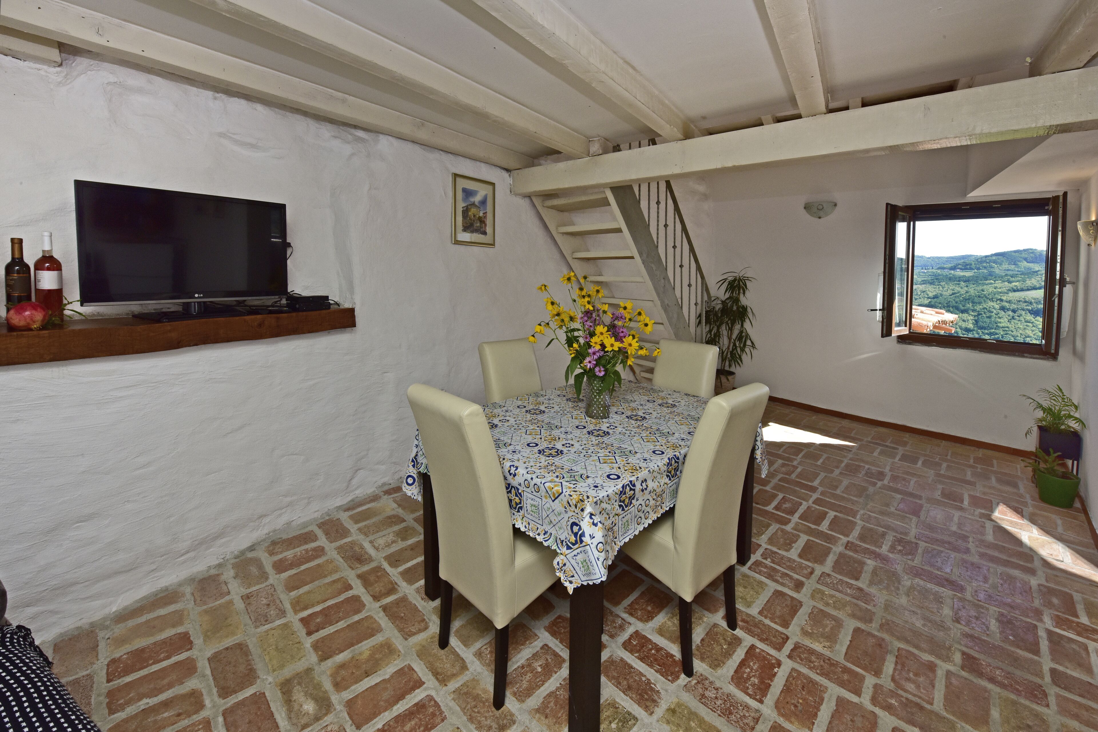 A dining area with a table set for four, a TV mounted on the wall, and a window with a view of the countryside.