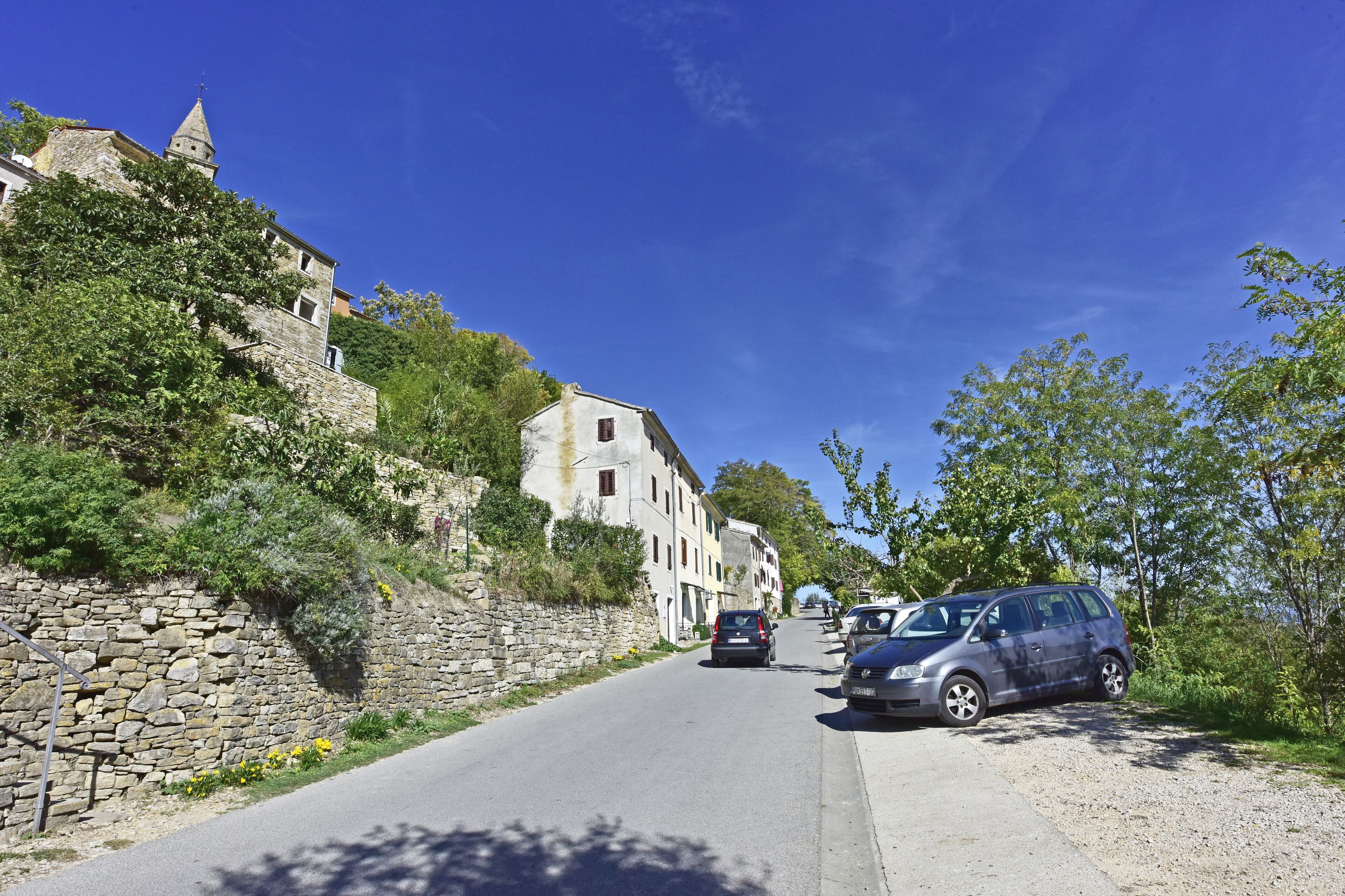 A narrow road flanked by stone walls and buildings, with parked cars and a clear blue sky.