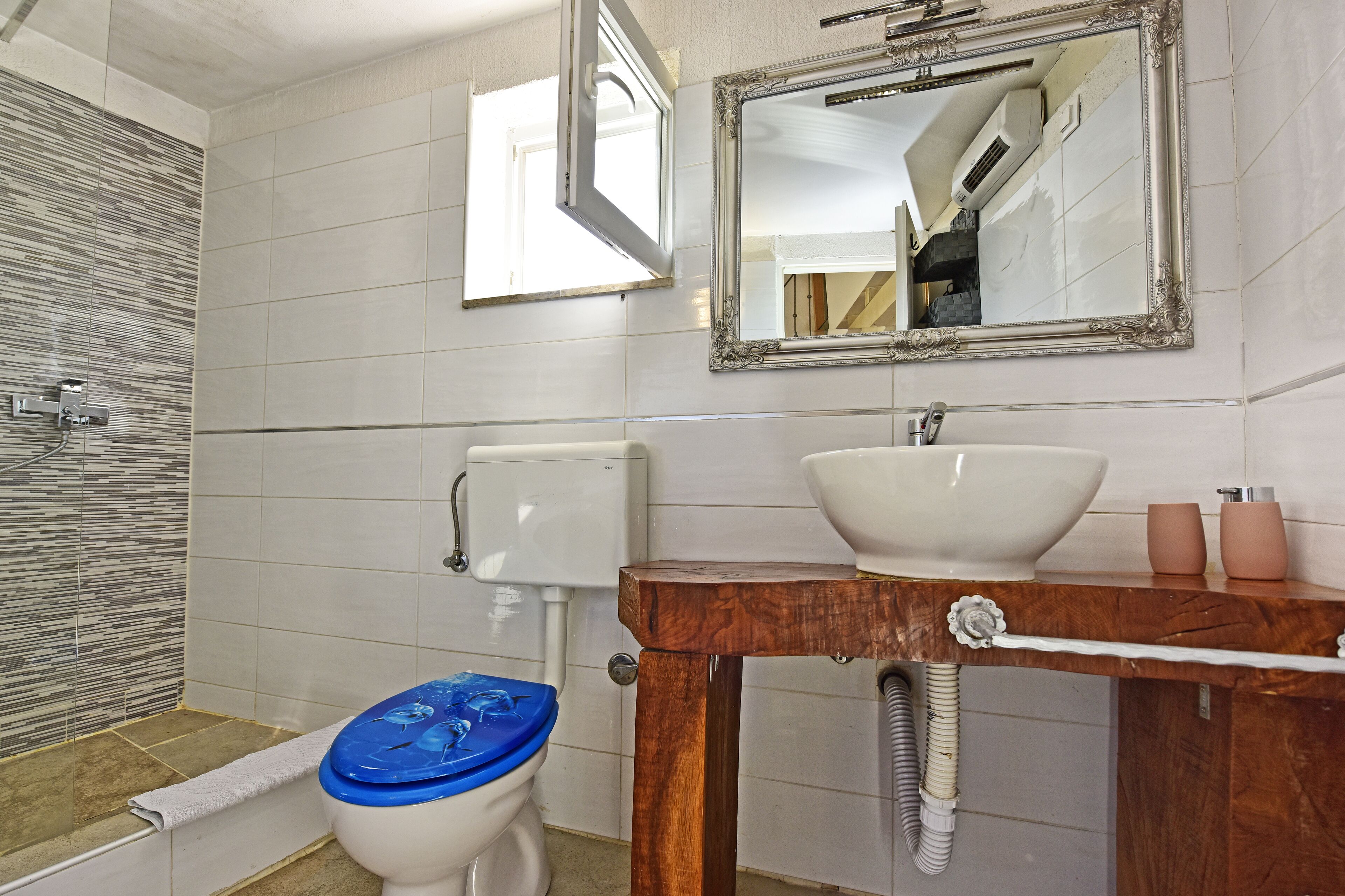 Une salle de bain avec des toilettes blanches, un meuble-lavabo en bois doté d’un grand évier et un miroir au-dessus du meuble-lavabo.
