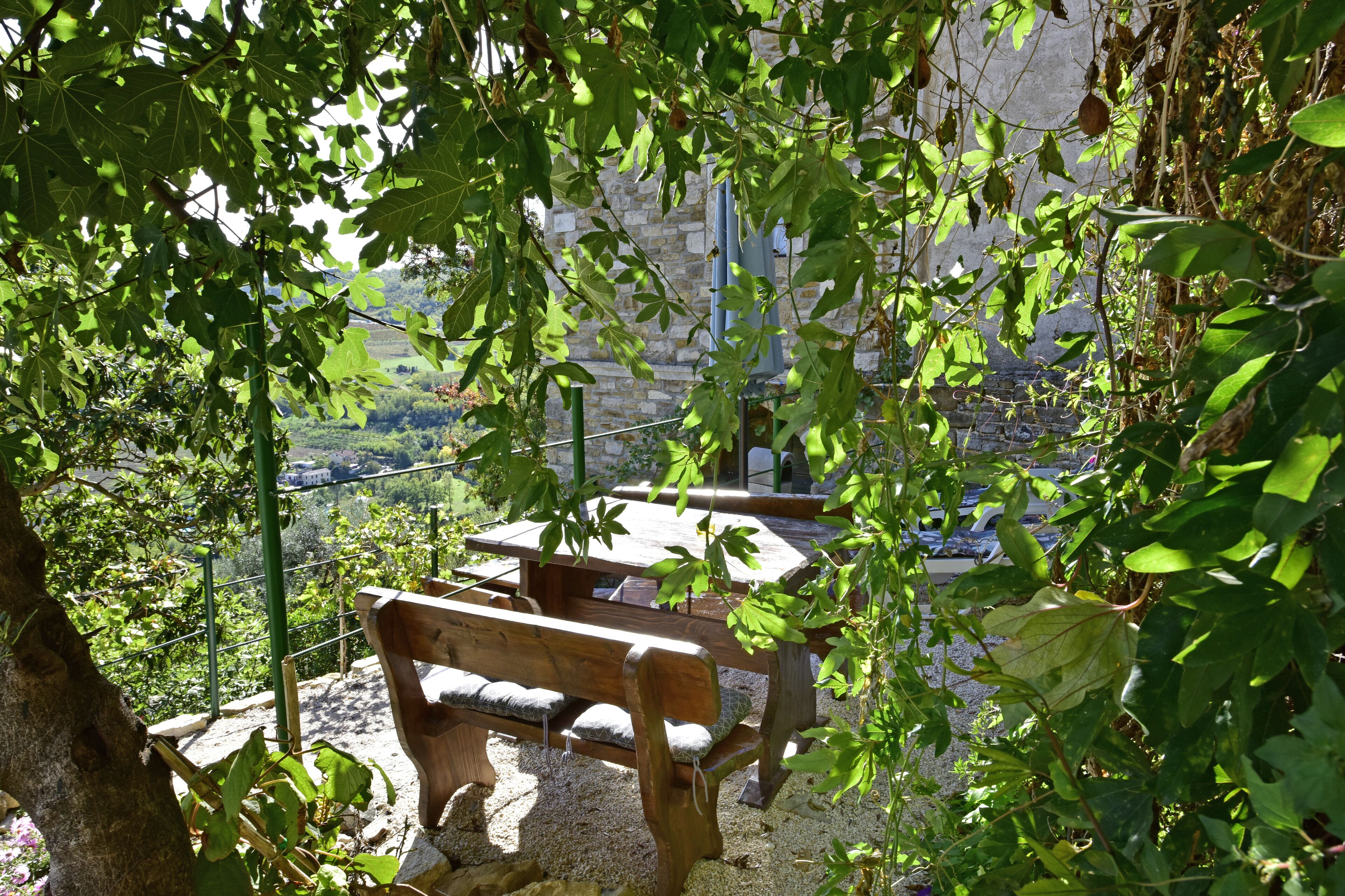 Outdoor wooden benches with cushions, surrounded by greenery and overlooking a stone building with a view of a valley.