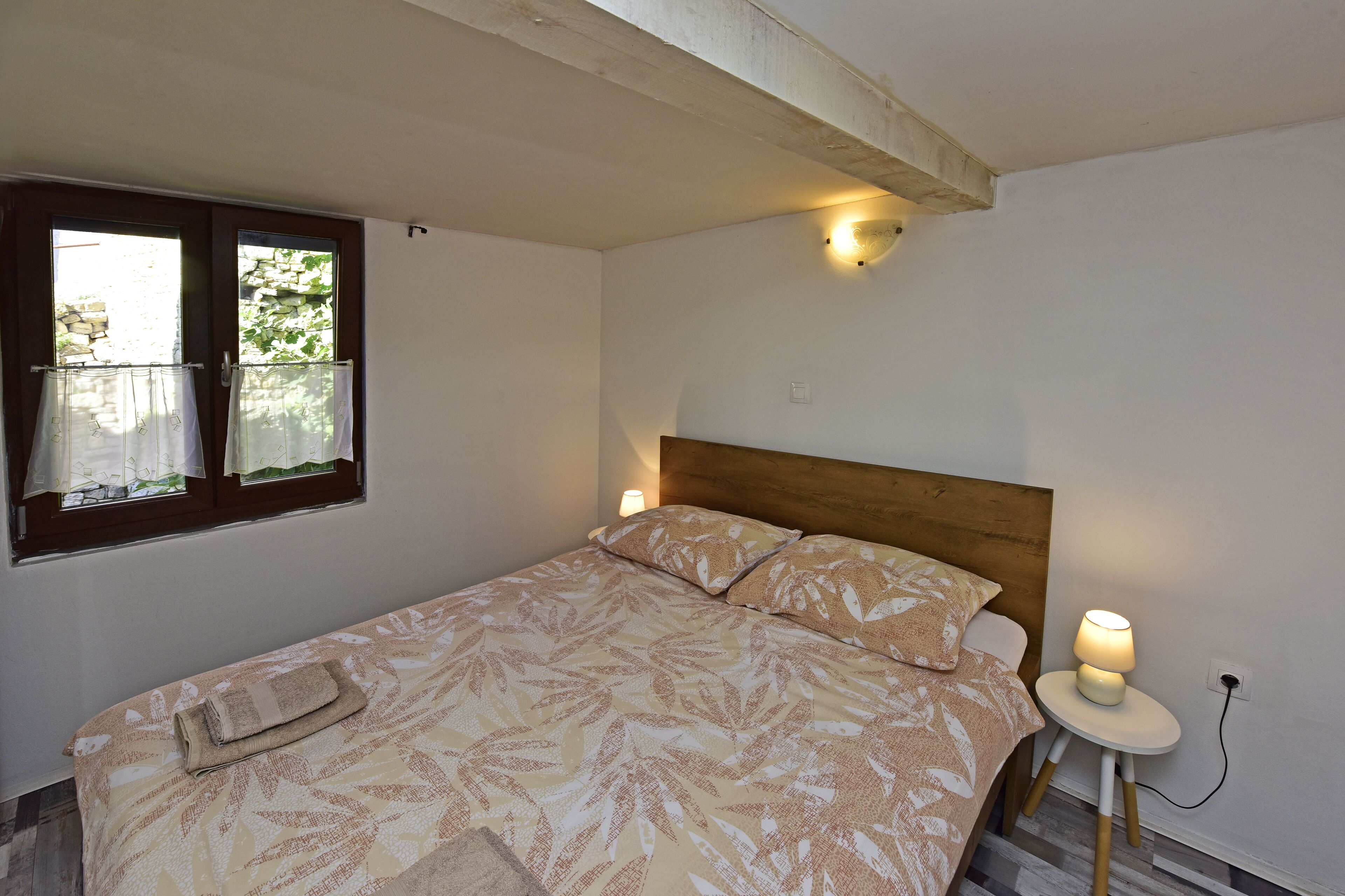 A bedroom with a bed, a nightstand, a lamp, a window with curtains, and a wall-mounted light fixture.