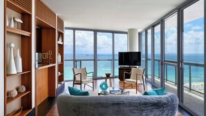 A spacious living area with a large sofa, two chairs, a coffee table, and a TV. The room features wooden shelving with decorative items and a large window offering a view of the ocean.