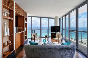 A spacious living room with a large gray sofa, two armchairs, a coffee table, and a TV. The room features wooden shelving with decorative items and a large window offering a view of the ocean. - Setai (Miami Beach)
