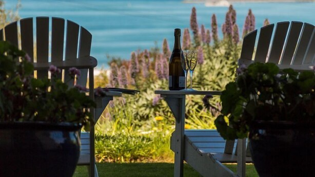 Two Adirondack chairs facing a table with a bottle and glasses, overlooking a coastal view with purple flowers and a grassy area. - Oceanfront Haven with Unparalleled Views and Retreat Living (Victor Harbor)