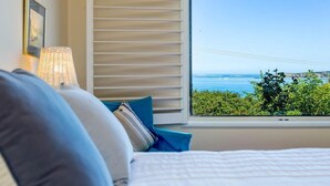A bedroom with a bed, a window with blinds, a lamp, and framed pictures on the wall. - Oceanfront Haven with Unparalleled Views and Retreat Living (Victor Harbor)