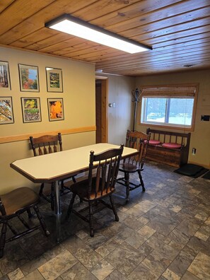 A dining area with a wooden table and chairs, framed pictures on the wall, a window with blinds, and a ceiling light. - Black Bear Lodge: Fall Colors on the Au Sable | Cozy Riverfront Cabin (Frederic)