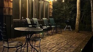A cozy outdoor patio with a wooden deck, a small building with large windows, and string lights. - Black Bear Lodge: Fish, Hike, and Relax in Michigan's Summer Paradise (Frederic)