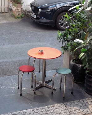 Un petit espace extérieur aménagé avec une table ronde et deux tabourets, un rouge et un vert, devant une voiture garée.