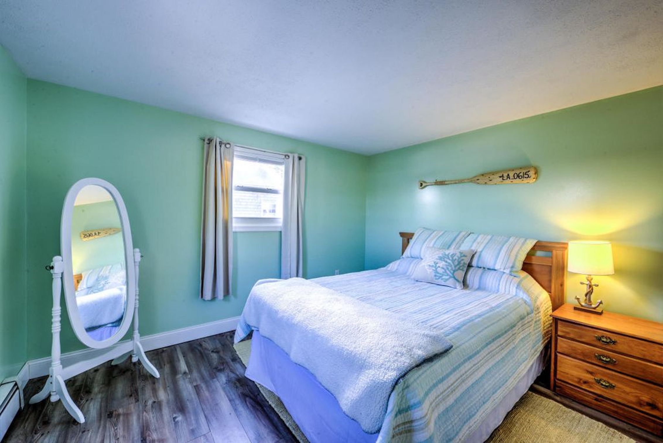 A bedroom with a bed, a vanity with a mirror, a window with curtains, and a nightstand with a lamp.