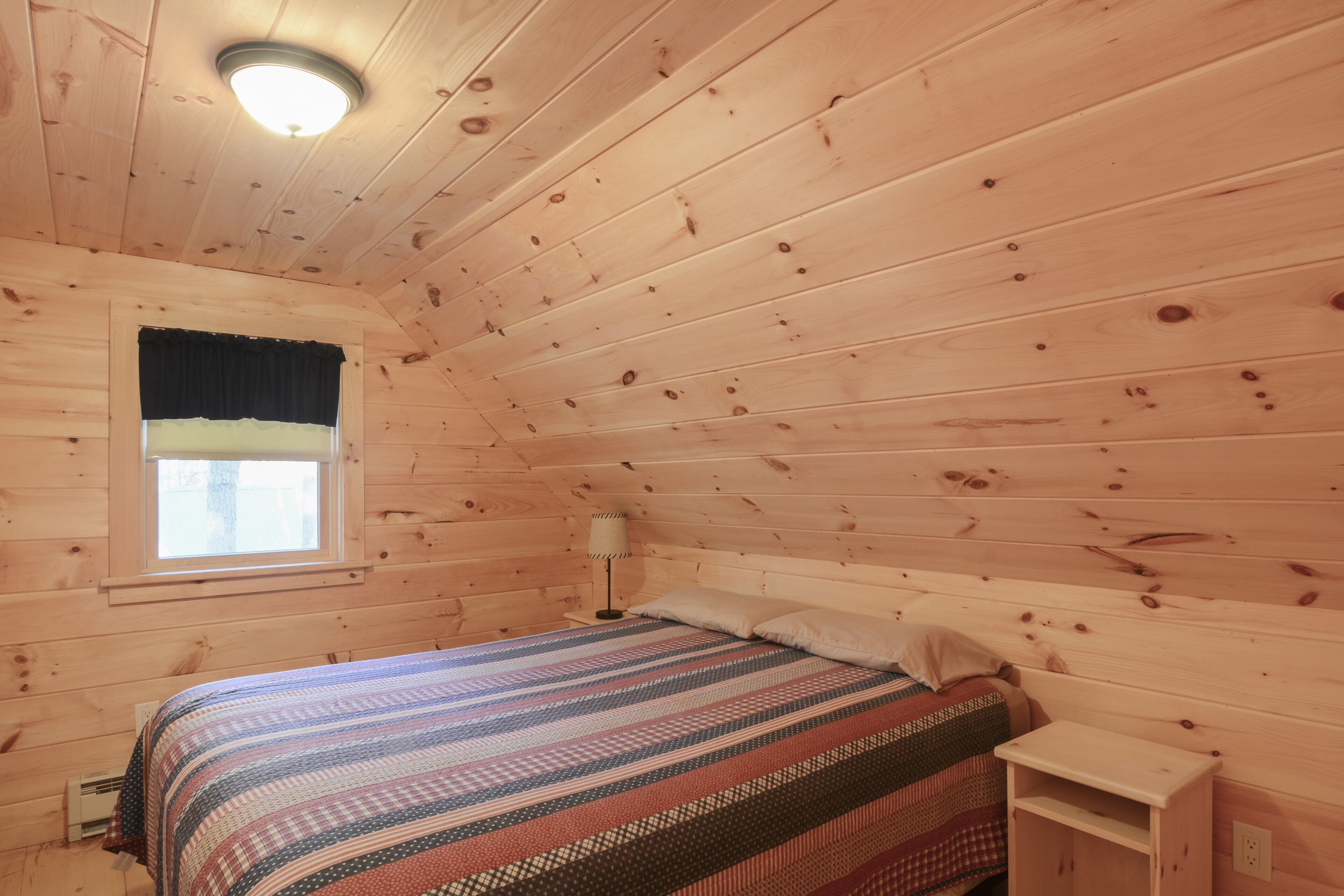 Habitación de cabaña de madera con cama, una ventana con cortina negra, una mesita de noche con lámpara y un artefacto de iluminación en el techo.