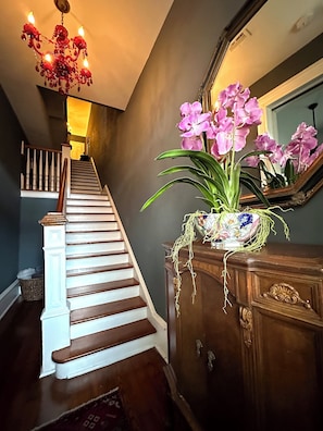 Un escalier avec des marches en bois et des balustrades blanches, un lustre à plusieurs lampes et un vase contenant des orchidées roses, posé sur une commode en bois.