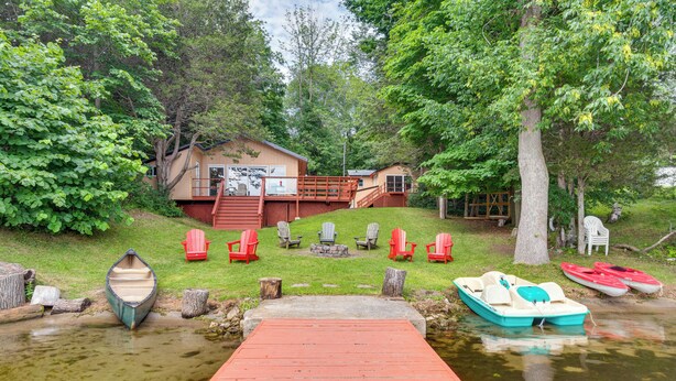 A lakeside cabin with a dock, red Adirondack chairs, and a small boat.