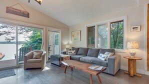 A spacious living room with a grey sofa, a beige armchair, and a wooden coffee table. There are large windows offering a view of the outdoors.