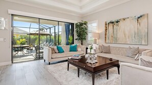 Un salón amplio con un gran ventanal corredera que da a un patio exterior, un sofá color beige con cojines verde azulado, una mesa de centro y un cuadro abstracto de gran tamaño en la pared.