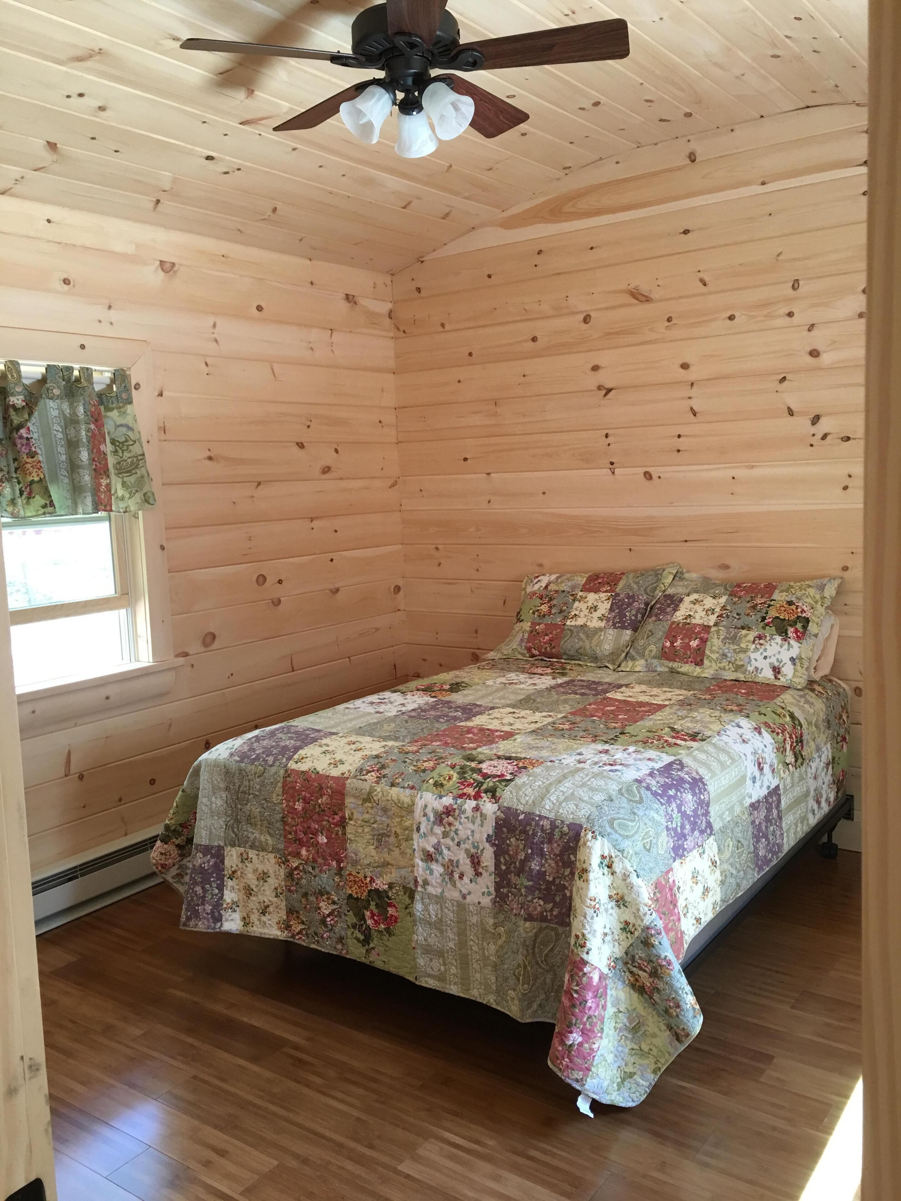 A cozy wooden cabin room with a bed, a ceiling fan, and a window with curtains.