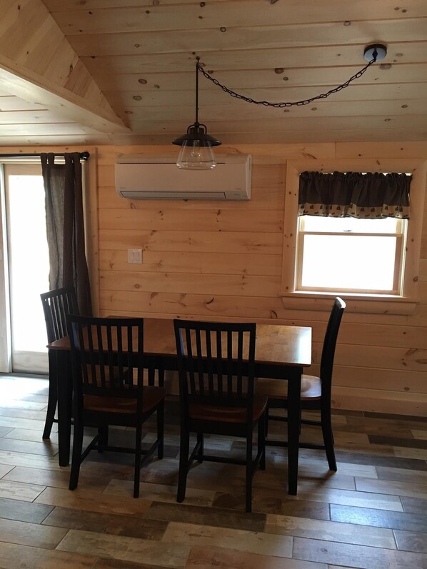 Un comedor con una mesa de madera y cuatro sillas, un equipo de aire acondicionado instalado en la pared y una ventana con cortinas.