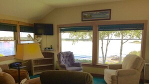 Un salon avec vue sur un lac, un téléviseur fixé au mur et une lampe sur une table d’appoint.