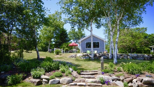 Une maison avec des fondations en pierre, entourée d’un jardin bien entretenu où poussent différentes espèces de plantes, et agrémentée d’un parasol rouge.