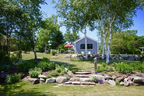 A house with a stone foundation, surrounded by a well-maintained garden with various plants and a red umbrella. - Breathtaking views, total relaxation, do as much or as little as you choose (Owls Head)