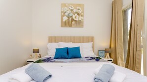 Ein ordentlich bezogenes Bett mit weißer und blauer Bettwäsche, ein Blumenbild an der Wand und ein Blick ins Freie durch beige Vorhänge.