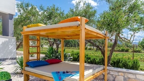 Ein hölzernes Bett mit Matratze und Kissen, auf einem Kiesgrund mit Garten und Bäumen im Hintergrund.
