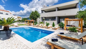 Ein Schwimmbecken mit Liegestühlen und einer Pergola, umgeben von weißen Kieselsteinen und viel Grün.