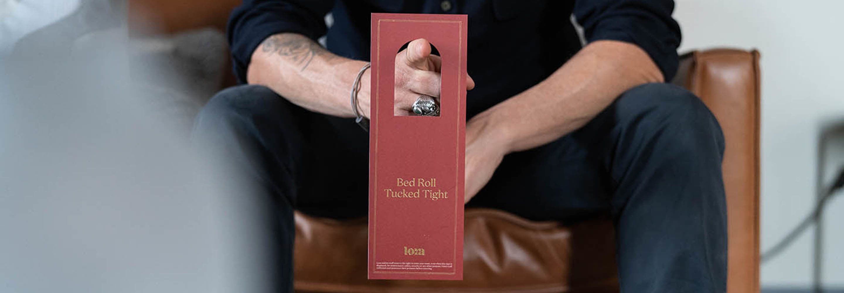 Una persona sentada en un sillón de cuero sosteniendo una caja roja con la etiqueta 'Bed Roll Tucked Tight'.