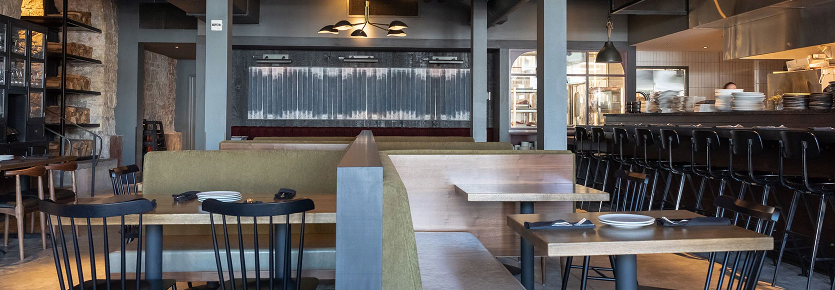 Un interior de restaurante moderno con mesas altas, cabinas, una zona de bar y vistas a la cocina.