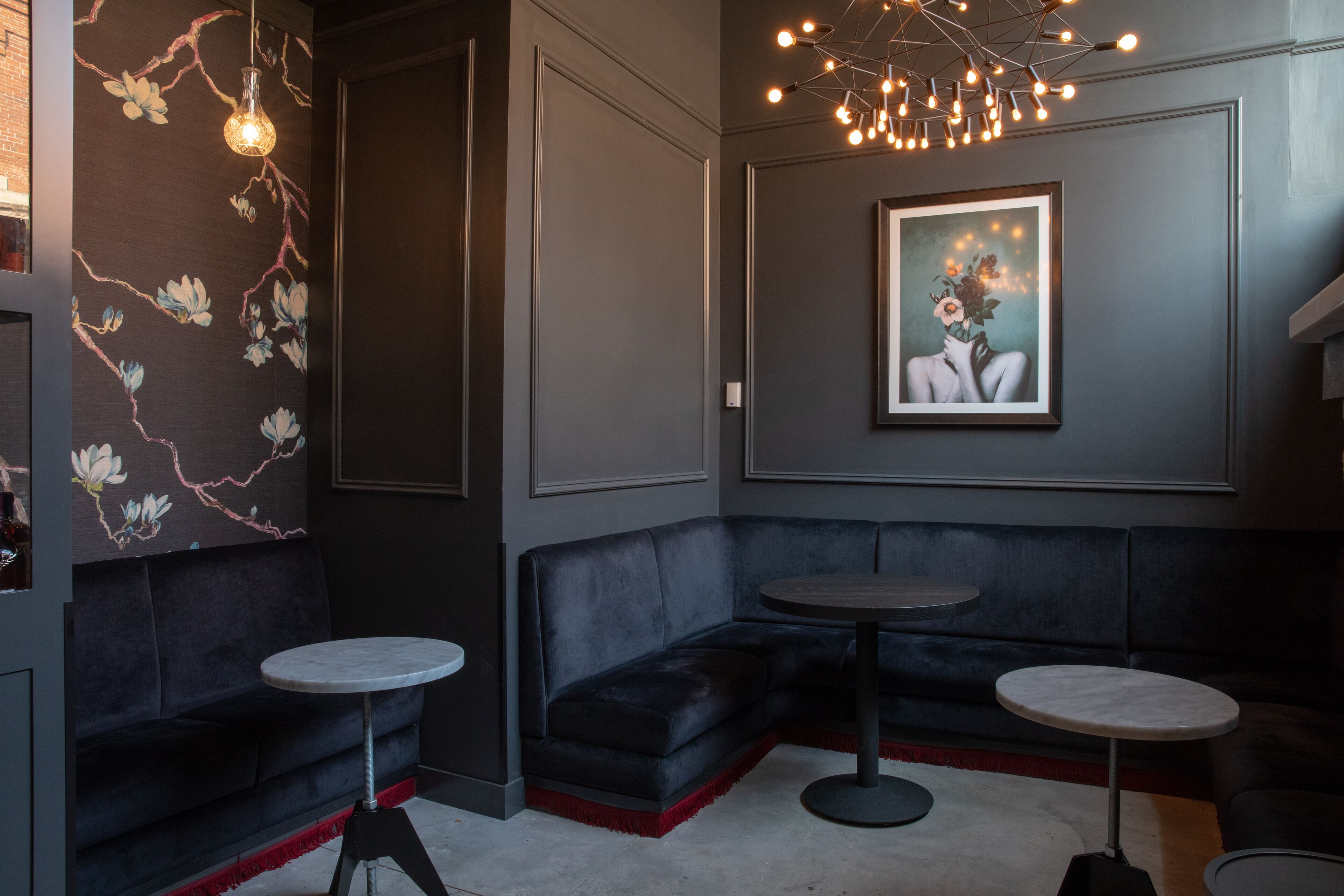 A modern lounge area with dark sofas, round tables, and a floral-patterned wall.