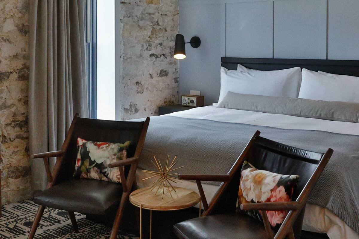A hotel room with a large bed, two chairs, a small table, a nightstand, and a wall-mounted lamp.
