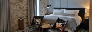 A hotel room with a large bed, two chairs, a small table, a nightstand, and a wall-mounted lamp. - Lora (Stillwater)