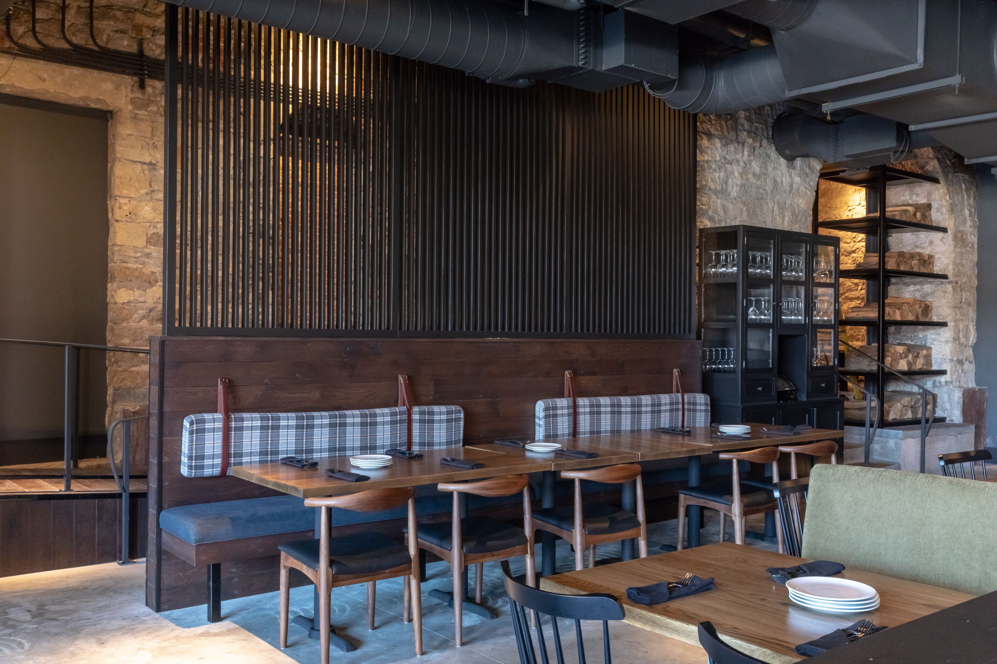 Un restaurant avec des tables et des bancs en bois, des murs en pierre et une étagère noire où sont rangés des verres.