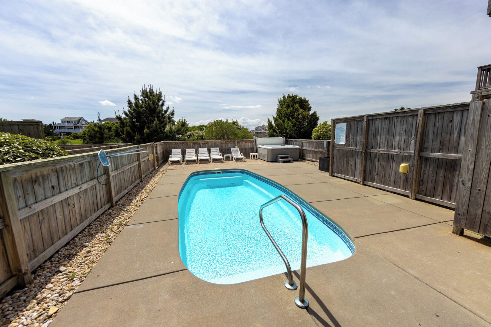 A rectangular swimming pool with a metal ladder, surrounded by a wooden fence, concrete deck, and several lounge chairs.