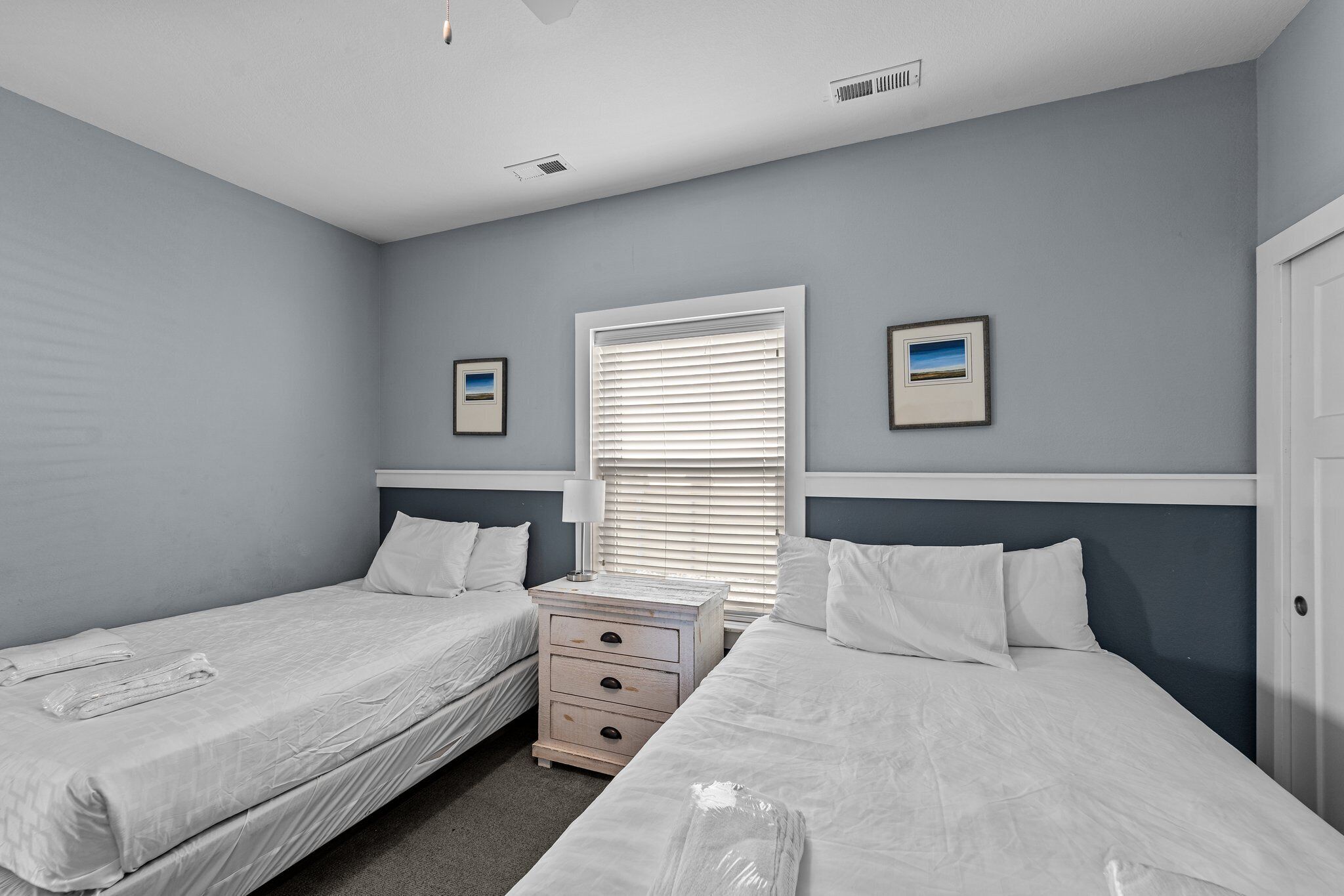 A room with two single beds, a nightstand with drawers, a window with blinds, and two framed pictures on the wall.