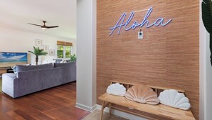 A living room with a grey sofa, a wooden accent wall with 'Aloha' neon sign, and a wooden bench with shell cushions. - 5 ★ Nakukui Hale: Waterfall Pool, Gazebo & Ocean Views, Gated Kona Town Retreat (Kailua-Kona)