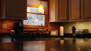Una cocina con armarios de madera, una ventana con persianas, un lámpara colgante, una cafetera, un tostador y un juego de cuchillos.