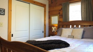 Un dormitorio con cabecera de madera, cama con ropa de cama blanca, manta de color negro, mesita de noche con lámpara y ventana con cortinas.