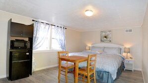 Un dormitorio con cama, lámparas de noche, una silla de madera, una mesita, microondas y refrigerador.